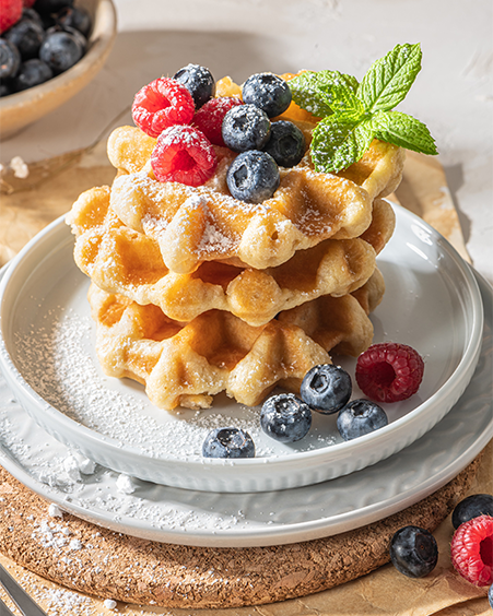 Stack of golden Belgian waffles dusted with powdered sugar, topped with blueberries, raspberries, lemon, and mint on a white plate